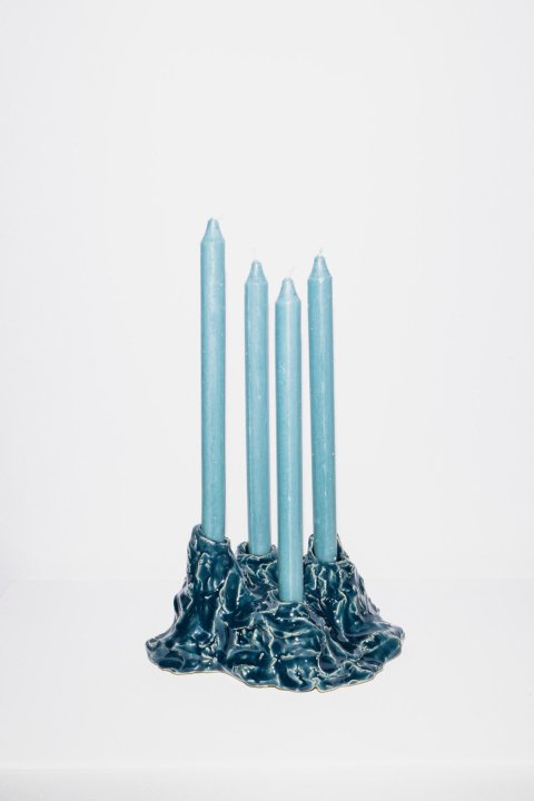 Ein blauer, handgeformter Kerzenständer aus glasiertem Ton mit vier eingesetzten blaufarbenen Stabkerzen, auf einem weissen Sockel vor einer weissen Wand.