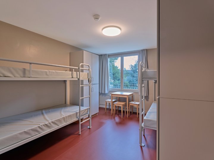 Schlicht eingerichtetes Mehrbettzimmer mit zwei Etagenbetten, weissem Schrank, kleinem Holztisch und Stühlen, sowie einem grossen Fenster mit Vorhängen.