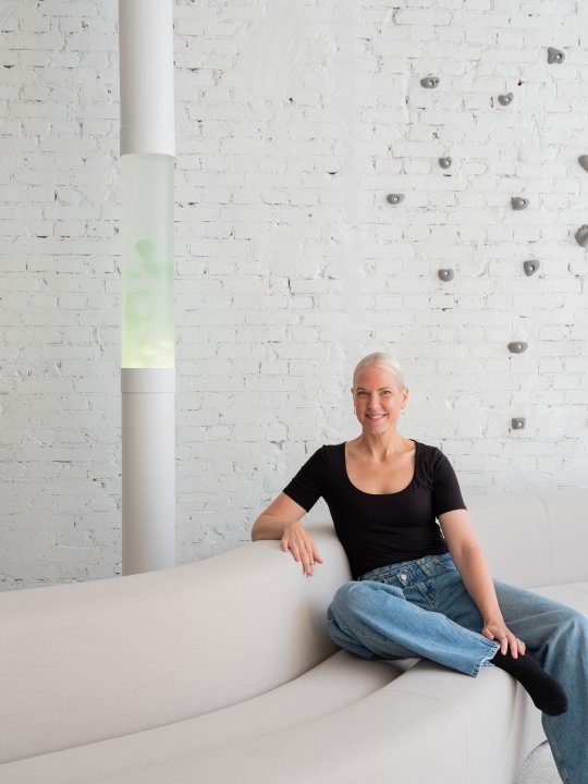 Die Designerin Sabine Marcelis mit kurzem blondem Haar sitzt auf einem beigen Sofa vor einer hohen Lavalampe mit grünlicher Lava und weisser Backsteinwand.