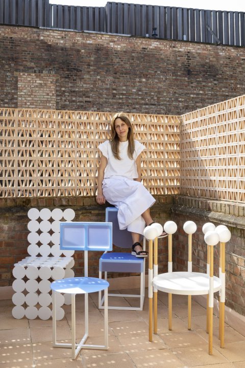 Designer Sara Leonor sitzt in ihrem Studio in London, umgeben von skulpturalen Stühlen in Blau, Weiss und Holzoptik.