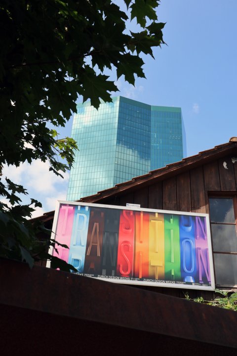 Plakat mit bunter Aufschrift Transition vor blauem Hochhaus und Holzfassade in Zürich.