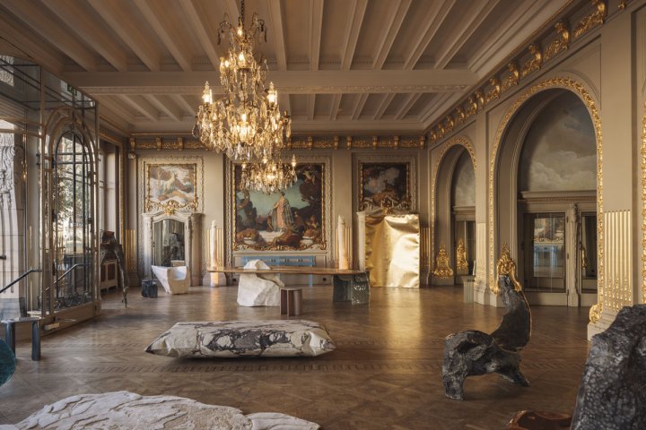 Ausstellungsraum mit zeitgenössischen Design- und Kunstobjekten in einem historischen Saal mit Kronleuchtern, Wandmalereien und ornamentalen Details.