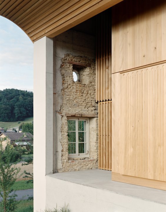 Längsseite eines alten Bauernhauses. Man sieht die geöffnete Fassade mit den alten Mauern und Fenstern und zurückversetzt im Innern einen Teil eines Holzkubus, der nachträglich als Wohnkörper integriert wurde.