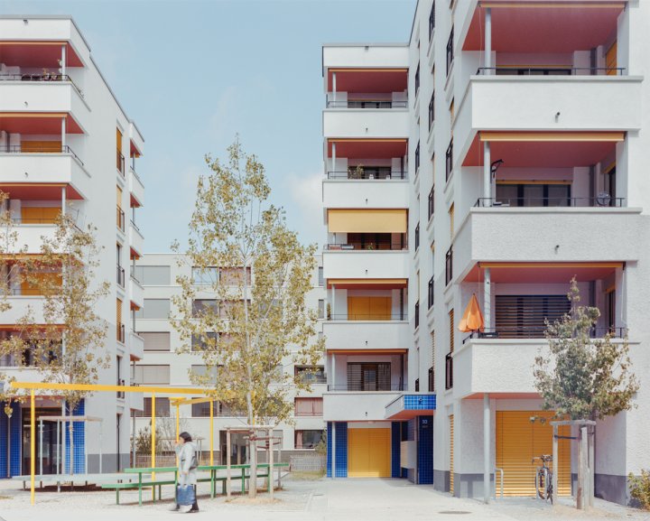Modernes Wohnensemble mit weissen Putzfassaden und farbigen Akzenten: Balkone mit rosafarbenen Decken, gelbe Storen und blau geflieste Eingänge; im Vordergrund ein Innenhof mit Bäumen und Sitzgelegenheiten.