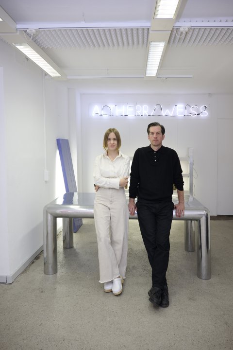 Ein Porträt des Zürcher Designduos Altherr Weiss, bestehend aus einer Frau und einem Mann, die in ihrem minimalistisch weissen Atelier vor einem markanten, glänzenden Metalltisch posieren. Die Frau trägt eine helle Bluse und weite helle Hosen, während der Mann in einem schwarzen Poloshirt und dunklen Hosen links neben ihr steht; im Hintergrund leuchtet ihr Name „ALTHERRAWEISS“ als weisse Neonschrift an der Wand.